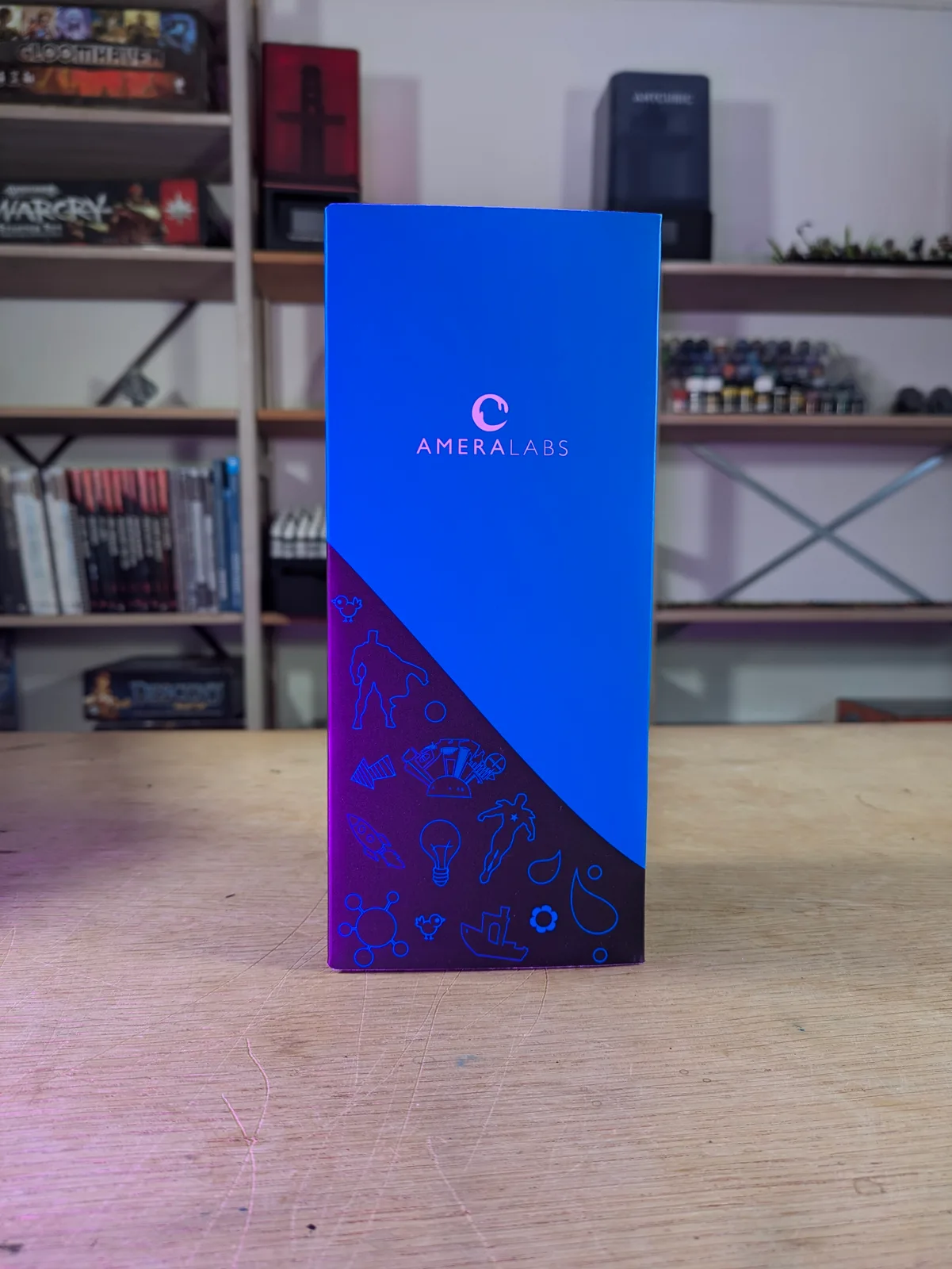 AmeraLabs retail box standing on a wooden workshop table with blue and dark purple illustrated packaging and AMERALABS branding, wargame bookshelves and an Anycubic printer visible behind