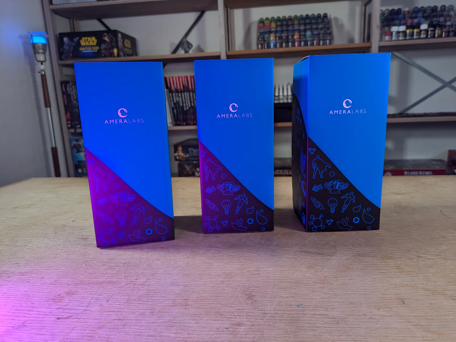 Three AmeraLabs retail boxes standing together on a workshop table with blue and dark purple gradient packaging, paint shelves visible behind