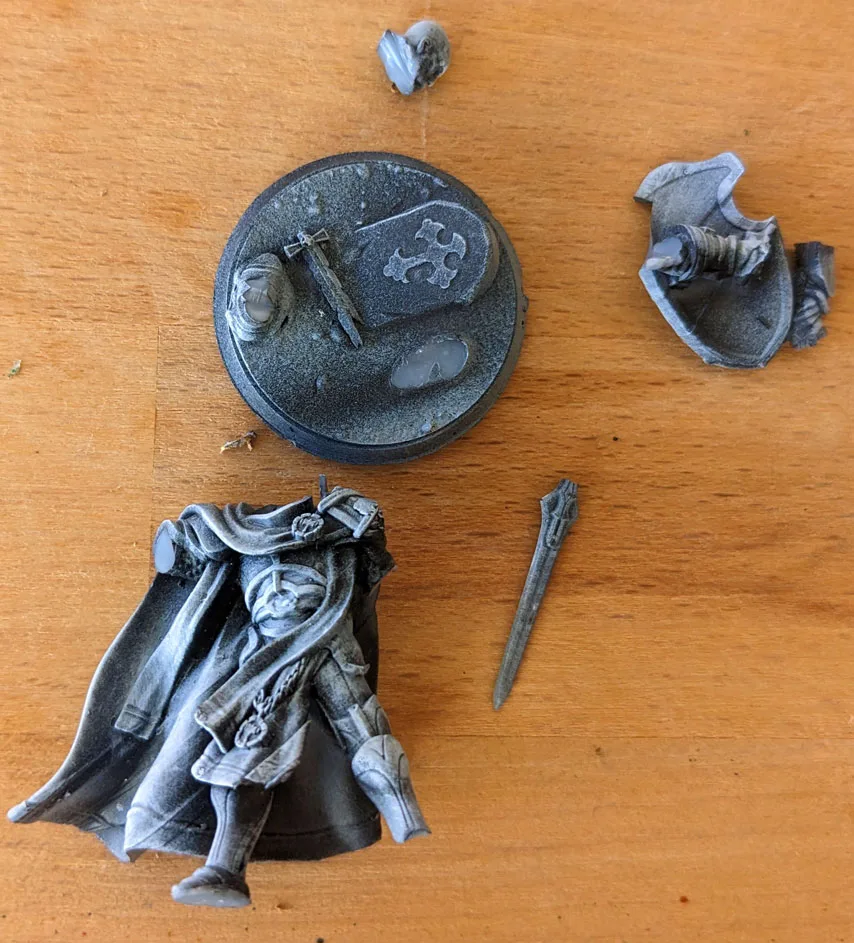 Black-primed grey resin cloaked warrior miniature shattered into five fragments on a wooden surface including lower torso with flowing cloak, round scenic base with shield, broken armoured arm, thin sword fragment, and small head shard