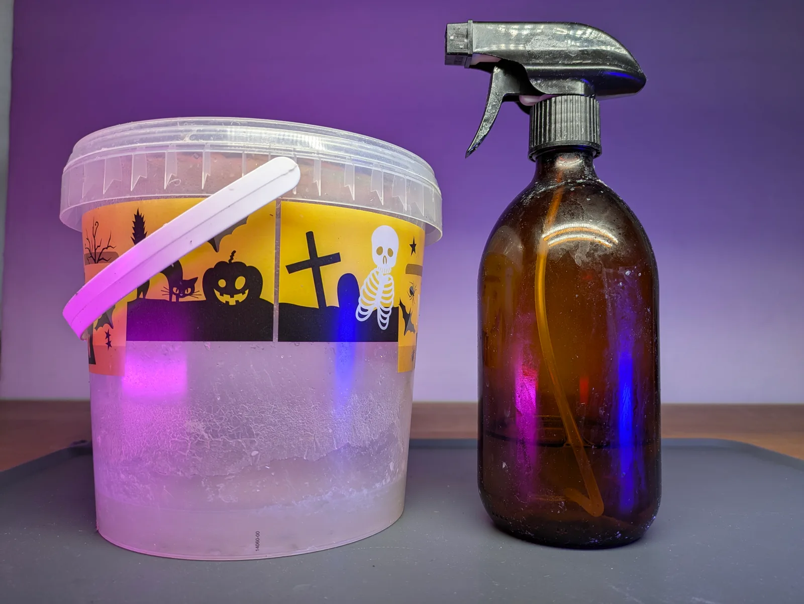 Clear plastic bucket with the lid ajar and a pink stirrer stick beside an amber glass trigger spray bottle on a grey mat, purple ambient glow