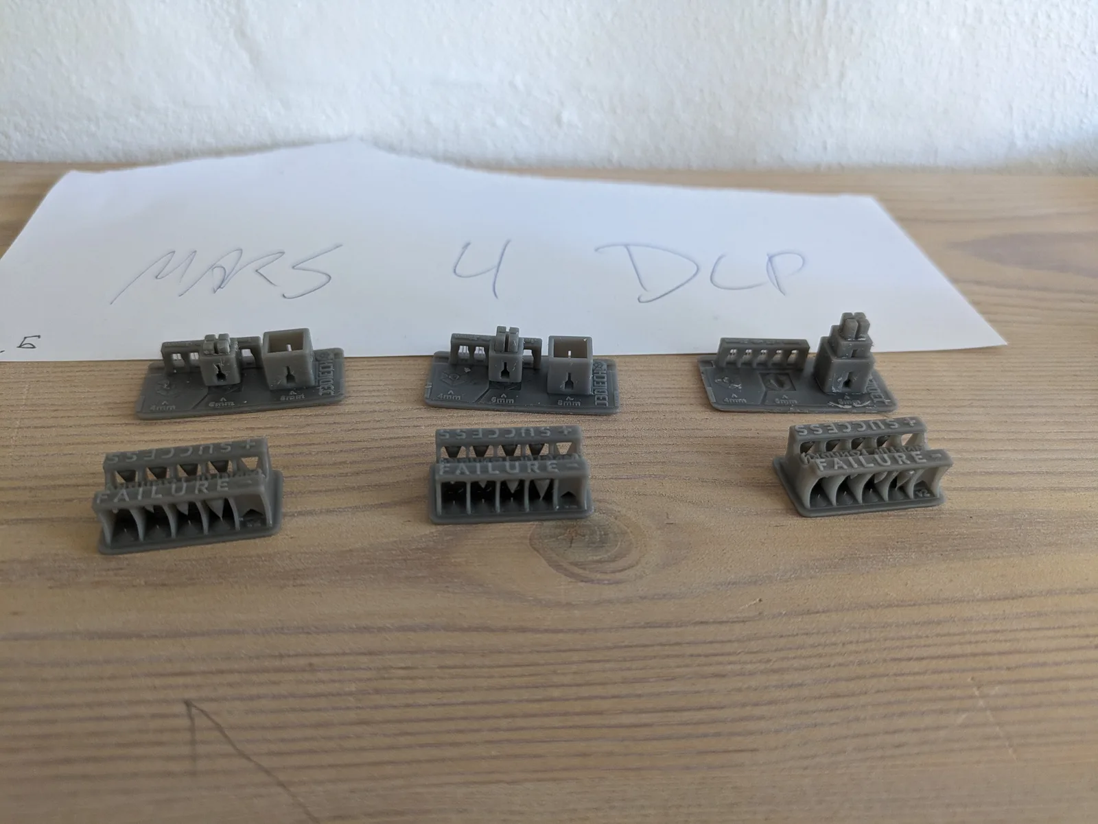 Three grey resin calibration sets in a row on a wooden table, each with a pin-tower bridge piece and a Cones of Calibration block with triangular teeth and SUCCESS plus FAILURE embossed text, with a handwritten MARS 4 DLP card behind