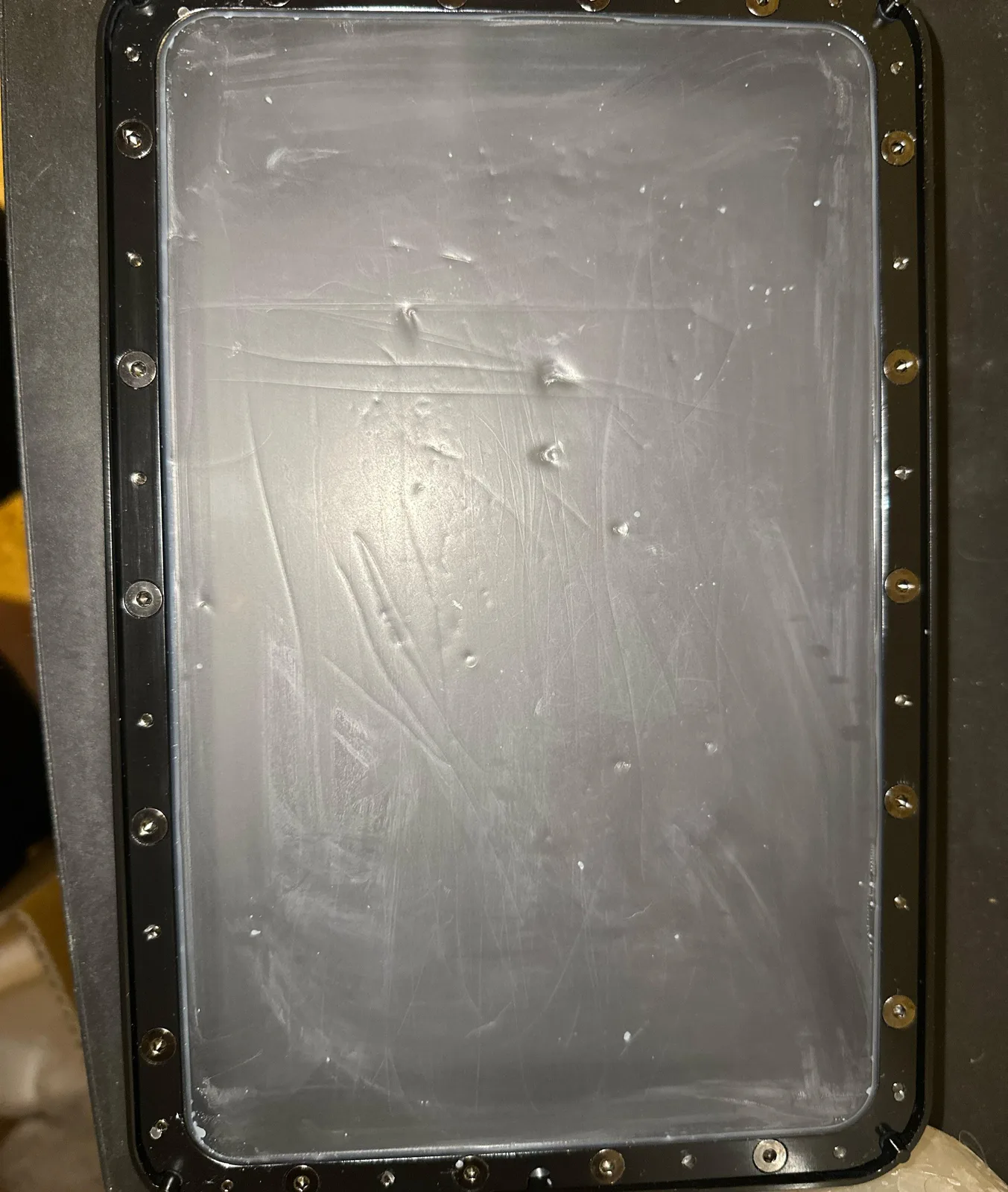 Close-up overhead of a resin printer FEP film showing heavy diagonal gouges, multiple small cloudy puncture holes, and milky white patches across the transparent membrane surface