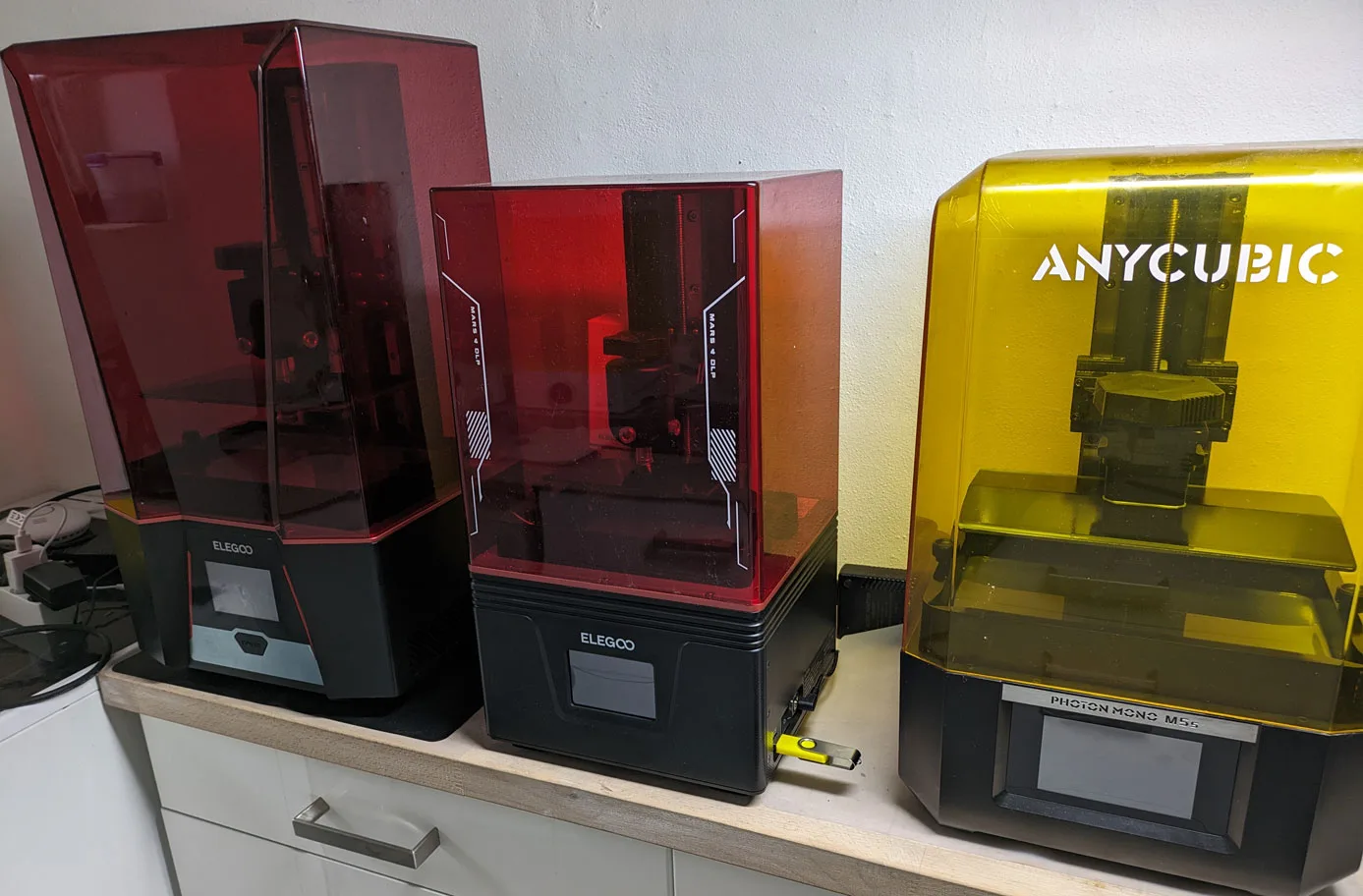 Two Elegoo resin printers with red acrylic lids and an Anycubic Photon Mono M5c with a yellow lid, lined up on a shelf