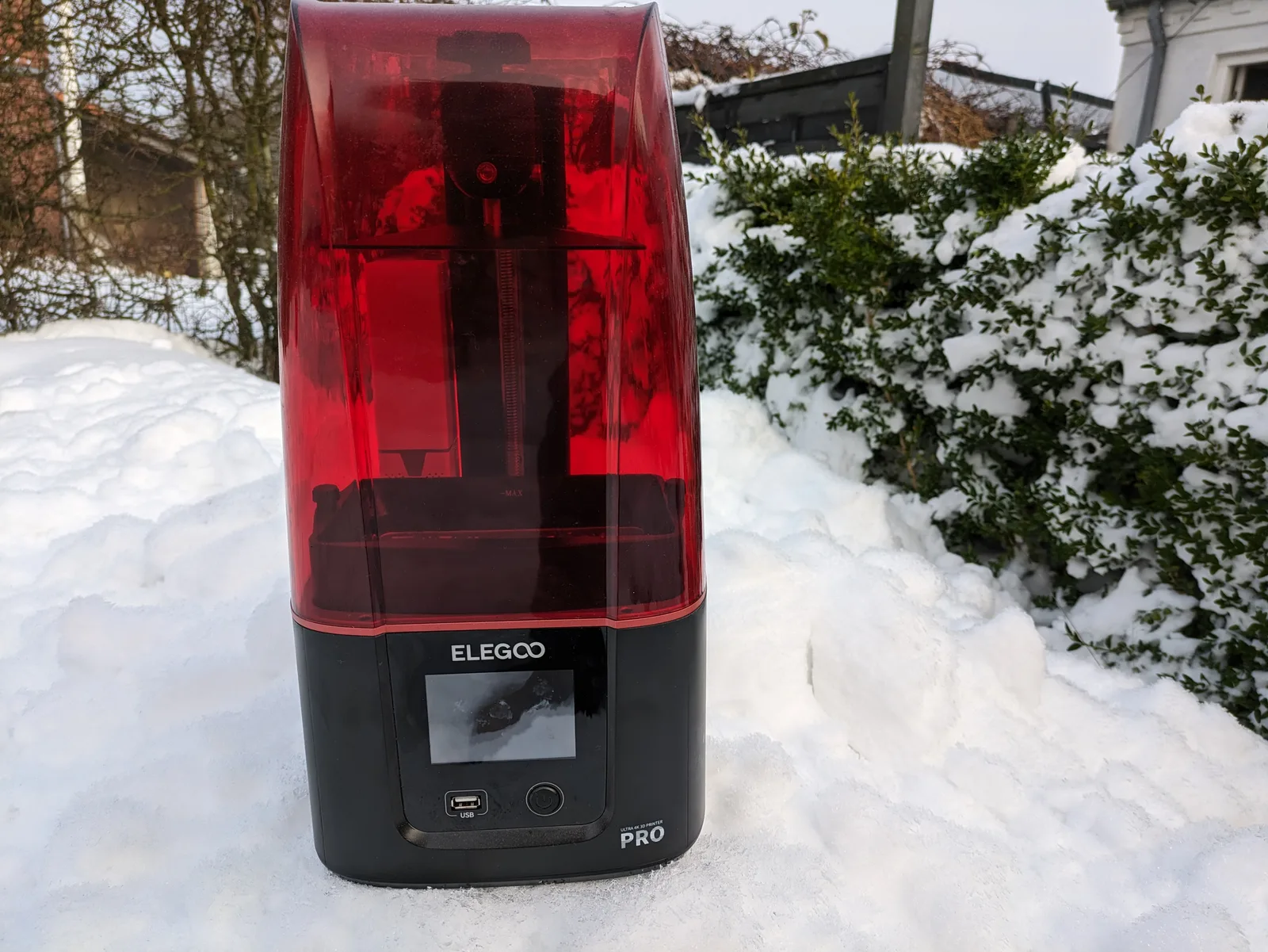 Elegoo Mars 3 Pro sitting on deep snow in a garden, red acrylic lid on, snowy hedge behind