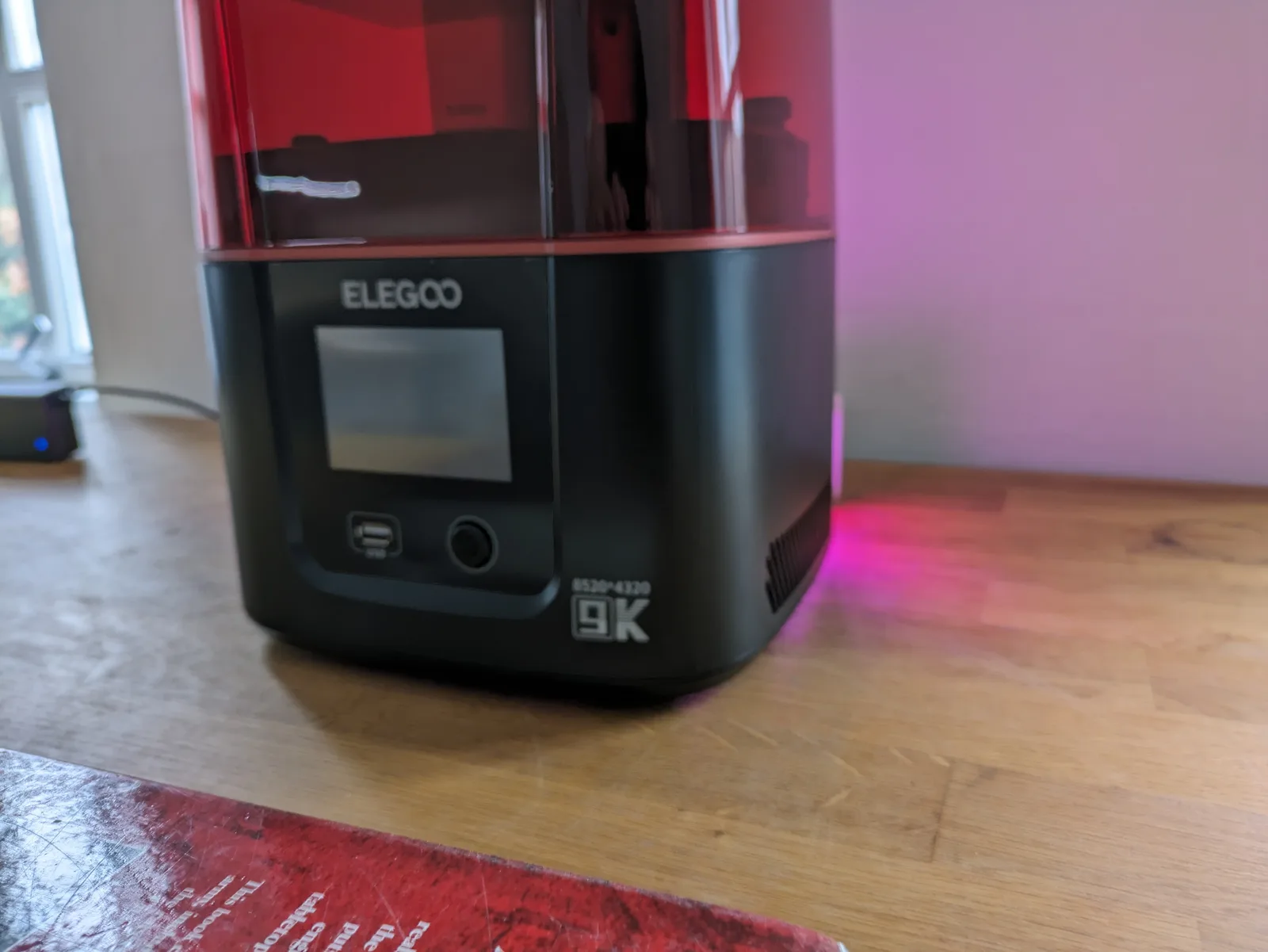Elegoo Mars 4 front panel: rounded red transparent lid with the 9K badge, idle touchscreen below, and a soft pink ambient LED glow