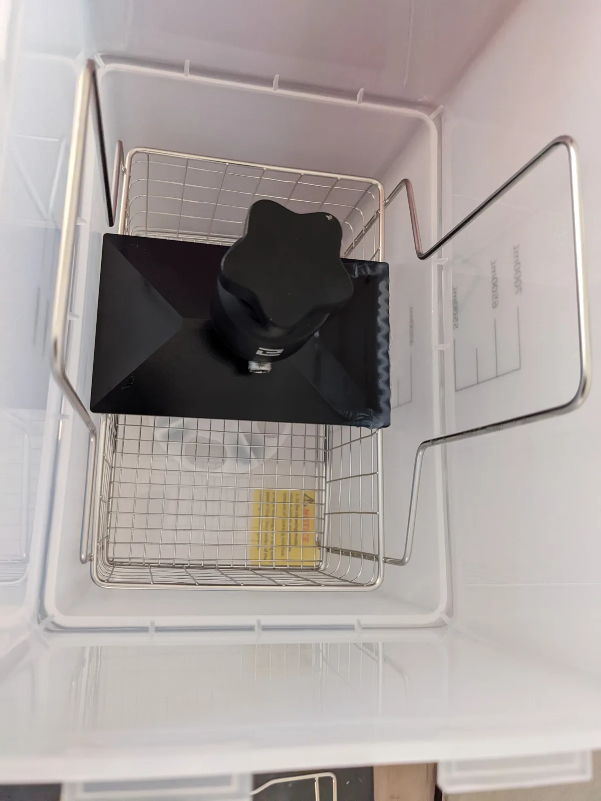 Top down view into an open Elegoo Mercury wash basin showing the stainless steel mesh basket with U shaped wire handle, a star shaped magnetic stirrer, and white interior walls with calibration level markings