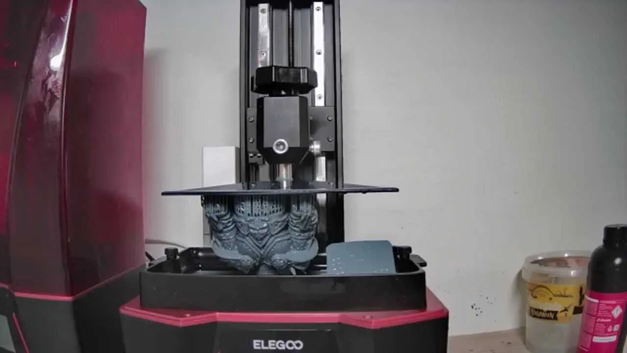 Elegoo Saturn printer hood removed showing build plate raised with a large dark grey complex resin print hanging from it, heavily detailed armoured figure or bust still attached to plate above the vat, Wargamer resin bottles visible to the right