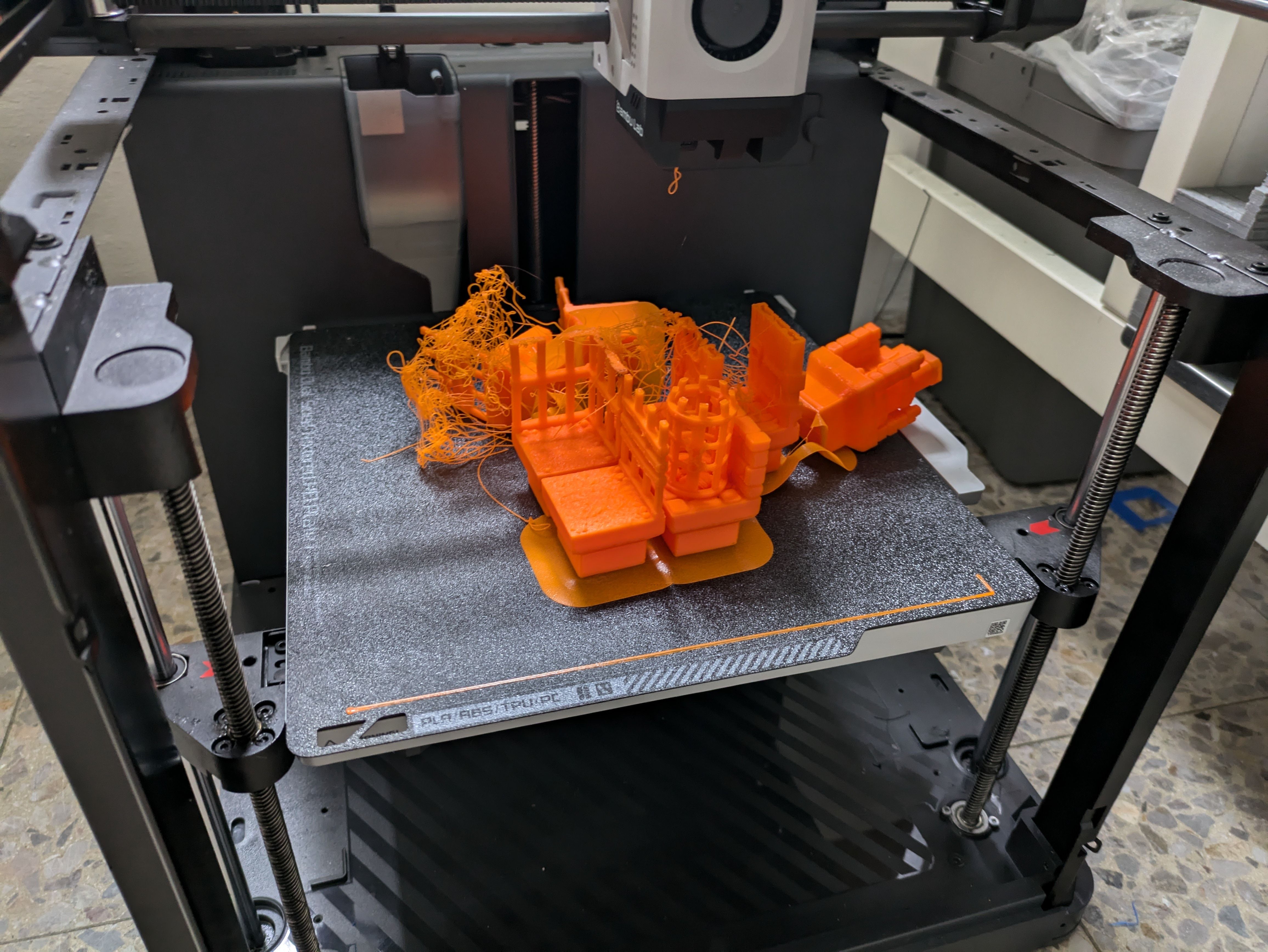 Catastrophic FDM print failure showing tangled orange PLA filament across the build plate, with several partially printed objects underneath