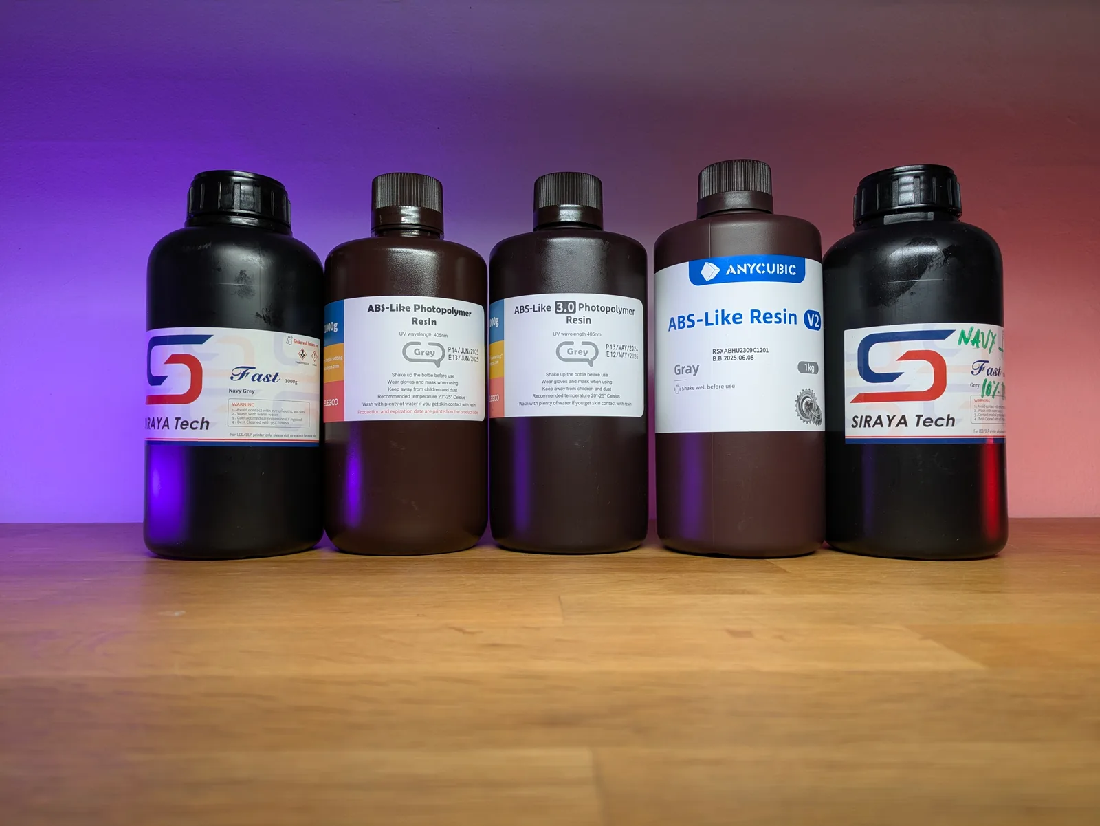 Five grey resin bottles in a row on a wooden surface: Siraya Tech Fast Navy Grey, two ABS Like Photopolymer bottles, Anycubic ABS Like V2 Grey and a partial Siraya Tech Fast