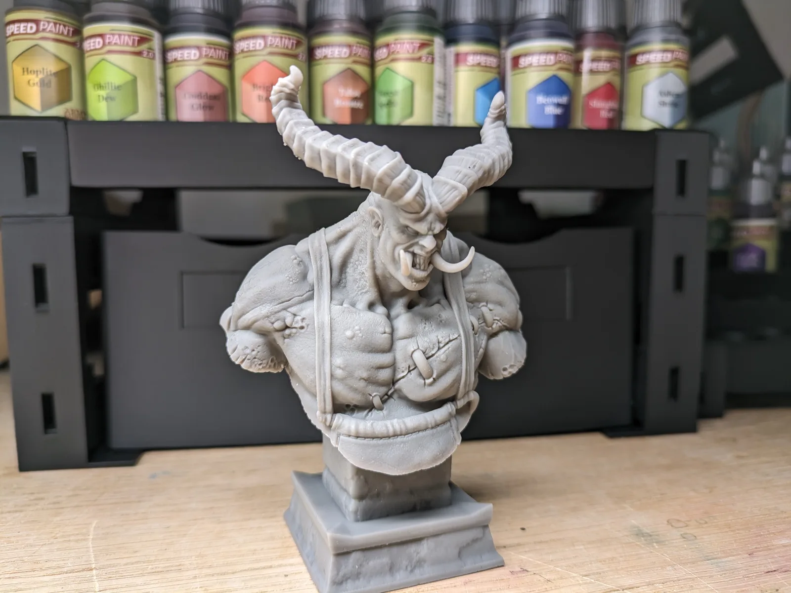 Large grey resin demonic bust on a square display plinth with massive spiralling bull-like horns, heavily muscled bare chest, tusk piercings, snarling expression, leather straps with studs, Army Painter Speed Paint row behind