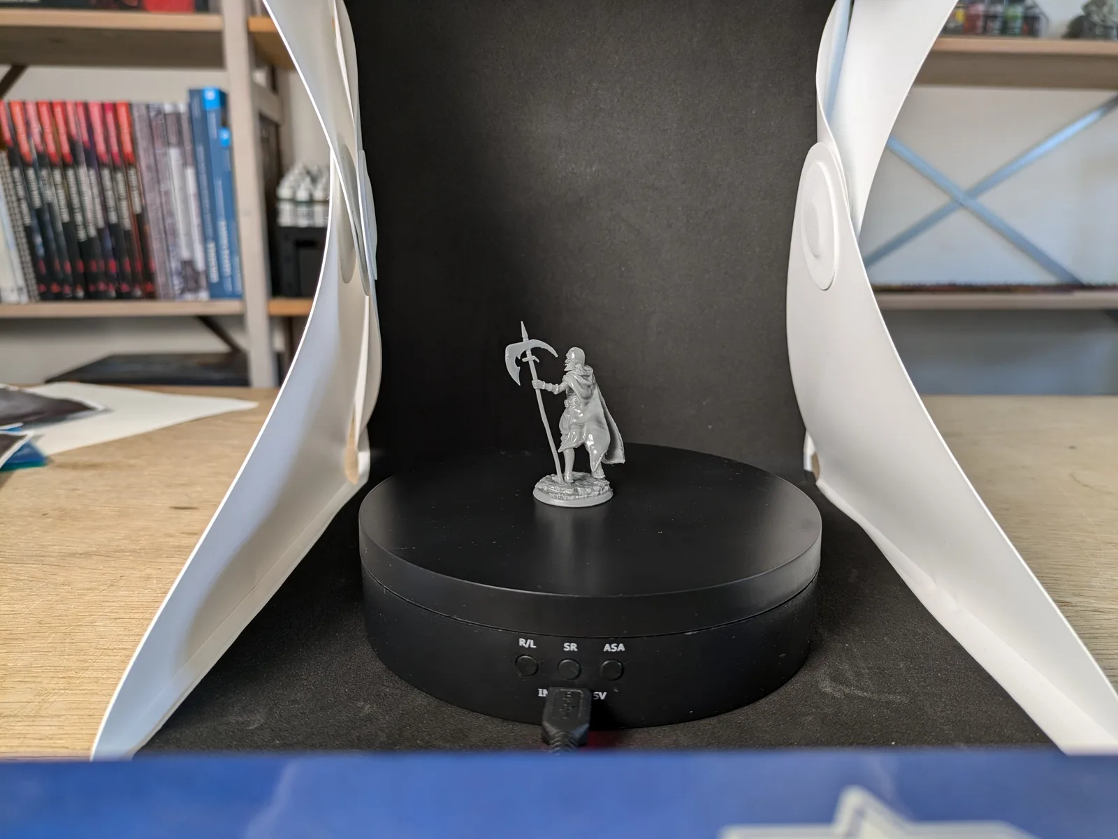 Grey resin undead armoured general holding a large scythe raised overhead, tattered cloak, dynamic stepping pose on rubble base, on a black rotating turntable in white and black lightbox