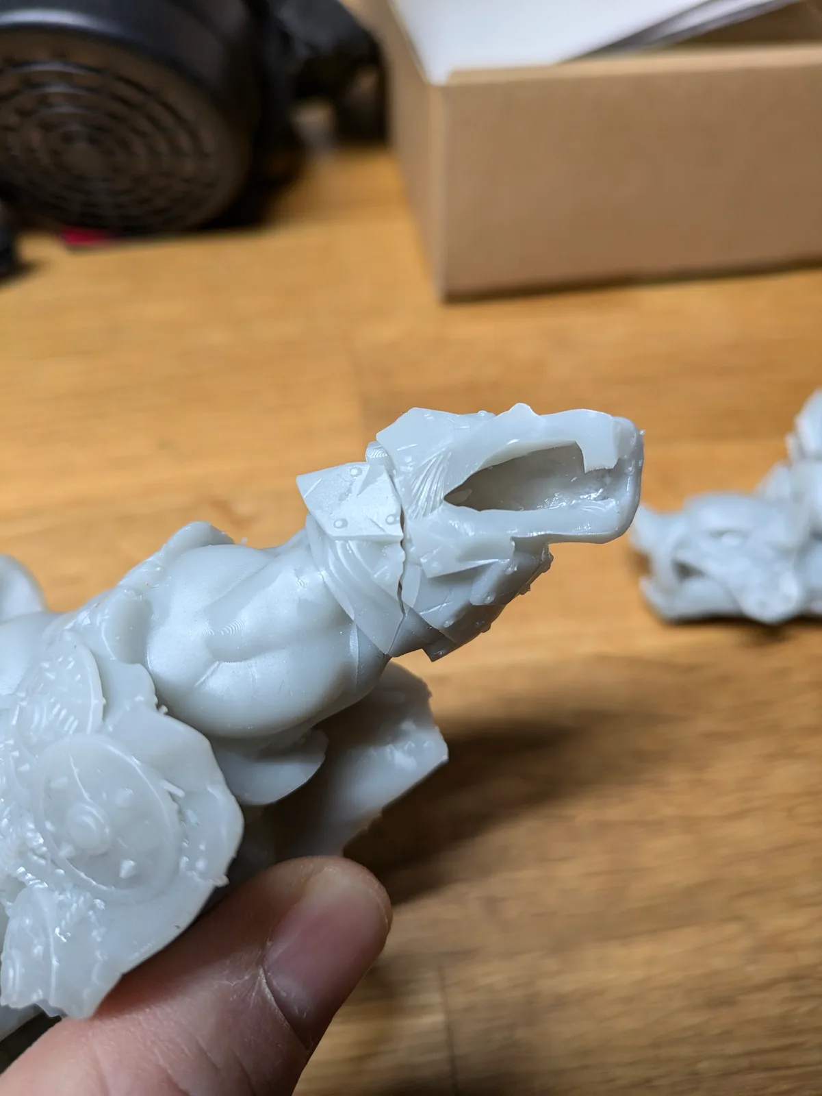 Hand holding a large white resin figure where the head and shoulder area has cracked open exposing the hollow interior, jagged delamination at the neck, a second cracked piece on the wooden surface behind