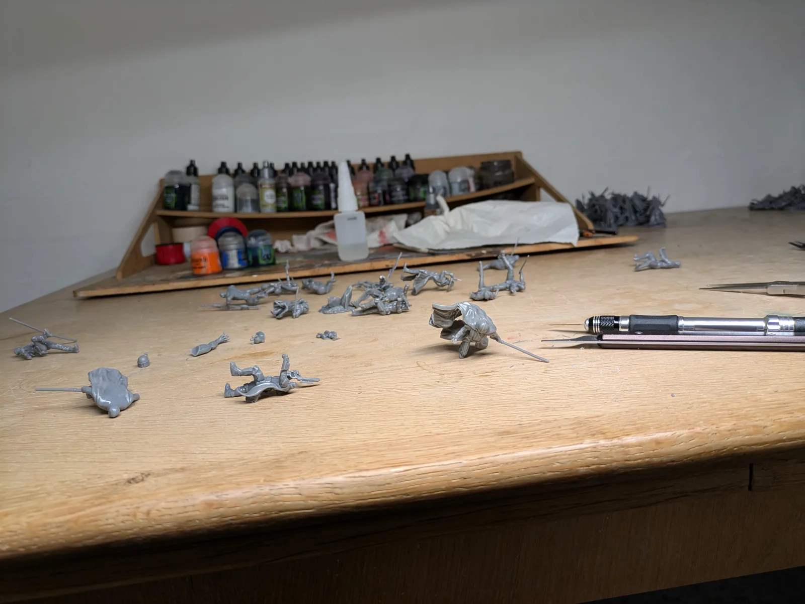 Hobby workbench covered with dozens of grey resin infantry miniatures after support removal, scattered arms and sprues, hobby tweezers at the right edge, paint bottles on the shelf behind