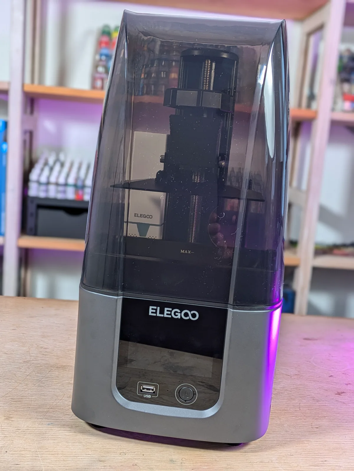 Full front view of the Elegoo Mars 4 Ultra with the smoked acrylic lid on, ELEGOO logo lit by UV LED, paint bottles on workshop shelves behind