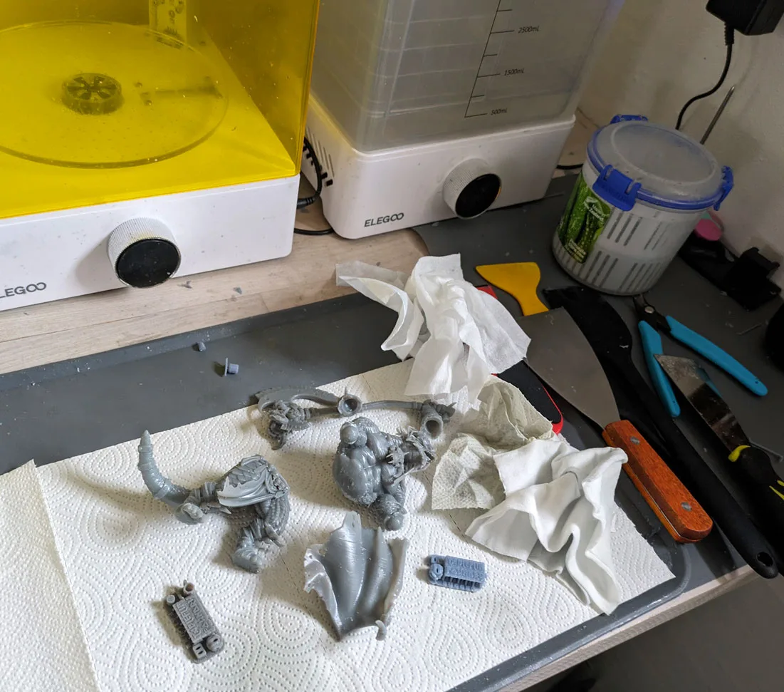 Two large grey resin fantasy miniatures on kitchen paper beside an Elegoo wash station, scraper and side cutters on the bench