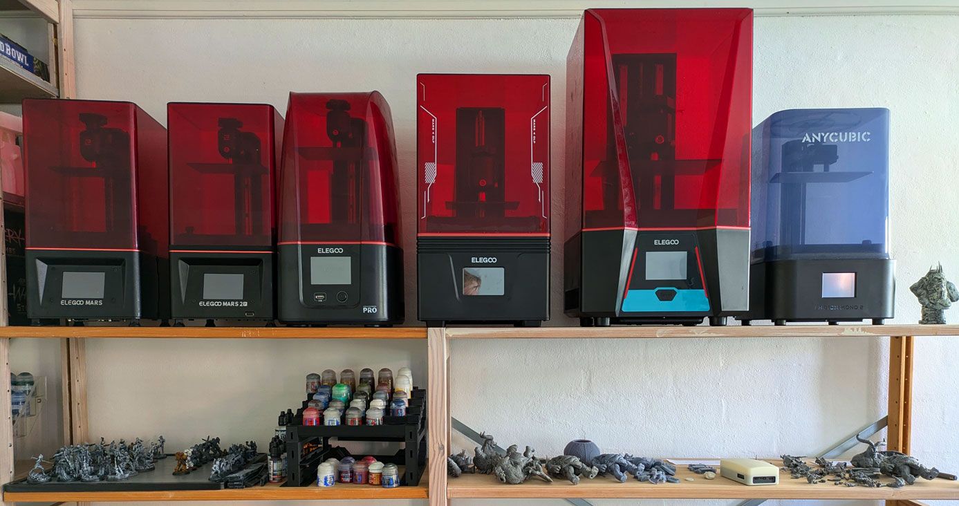 Row of Elegoo Mars and Anycubic resin printers on workshop shelves, paint pots and printed grey miniatures on the shelf below