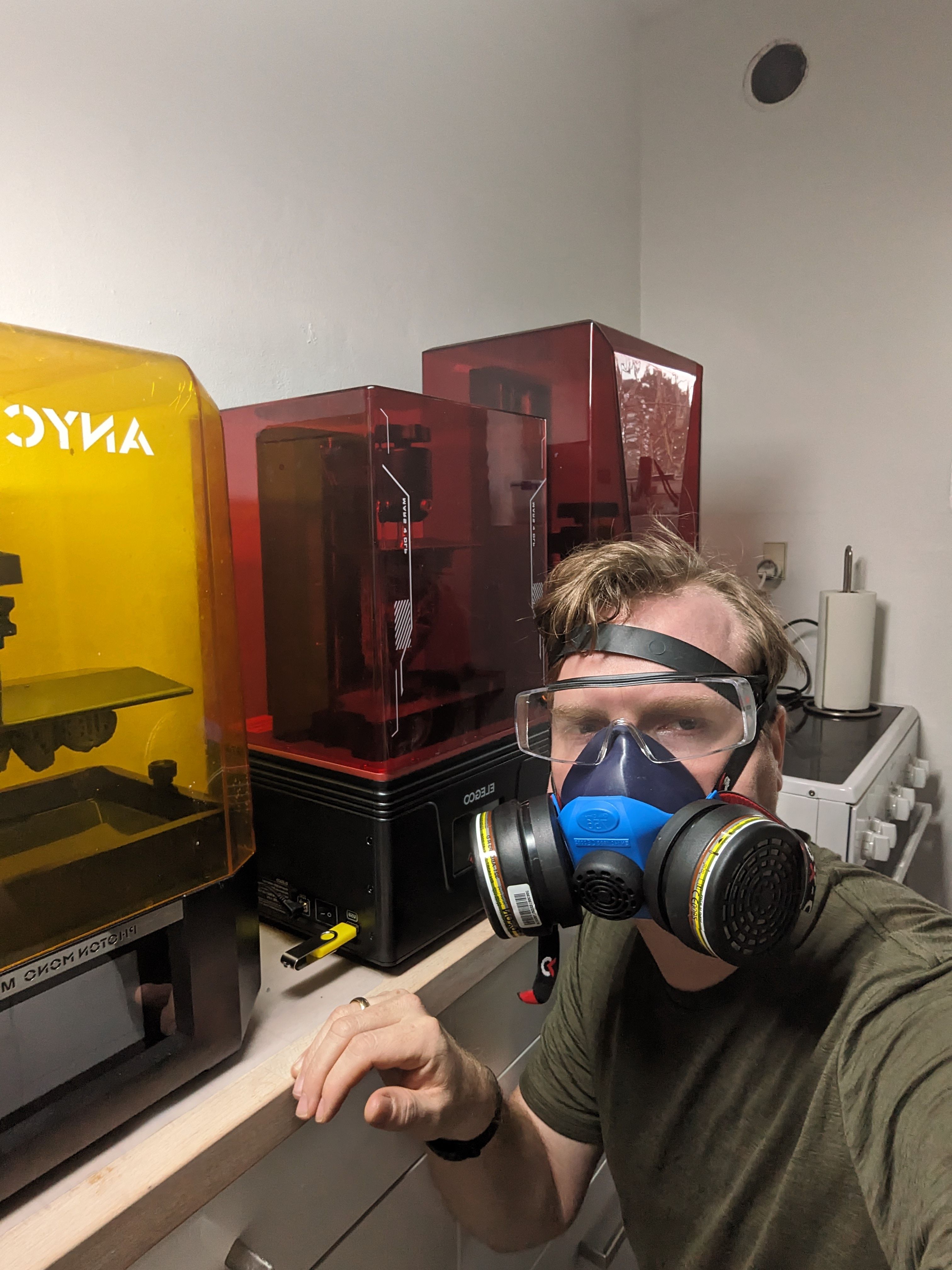 Peter Thuborg in a respirator and wraparound safety goggles standing in front of three resin printers in his dedicated printer room