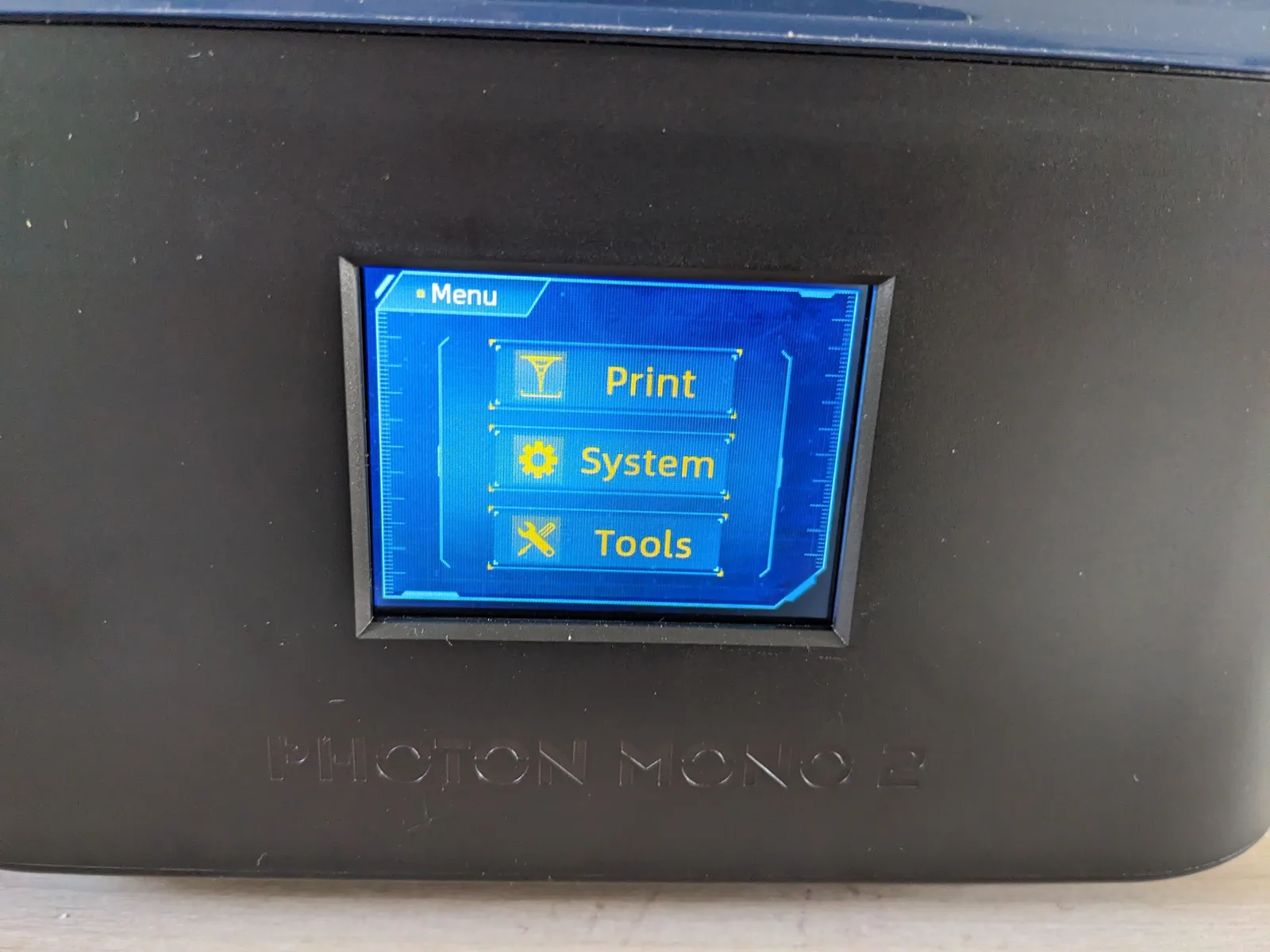 Touchscreen on the front of the Anycubic Photon Mono 2 lid showing the main menu with Print, System, and Tools options in orange on blue, with the embossed PHOTON MONO 2 label visible below