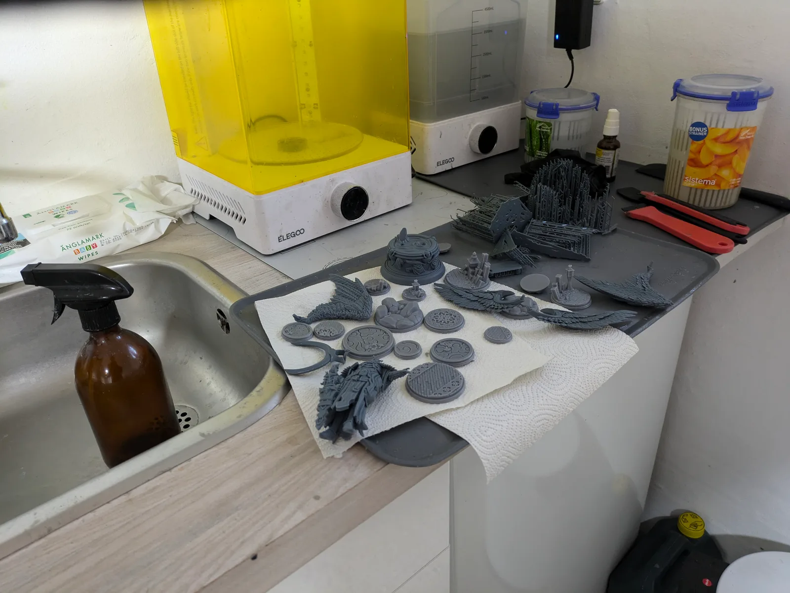 Workbench beside a kitchen sink with a yellow Elegoo Mercury Plus wash unit and a white Elegoo Mercury cure station on the worktop, grey resin miniatures and ornate bases drying on kitchen paper, an amber spray bottle in the sink