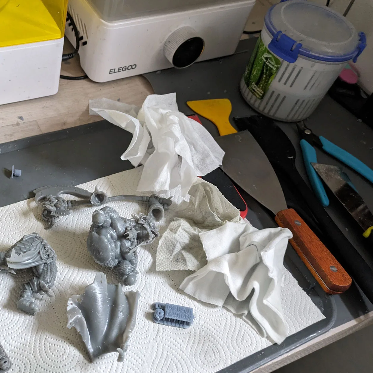 Post-wash workflow on a dark workbench mat, grey resin miniatures resting on paper towel, Elegoo Mercury wash station lid at top-left, yellow scraper and blue flush cutters laid out at the right, cure station in the background