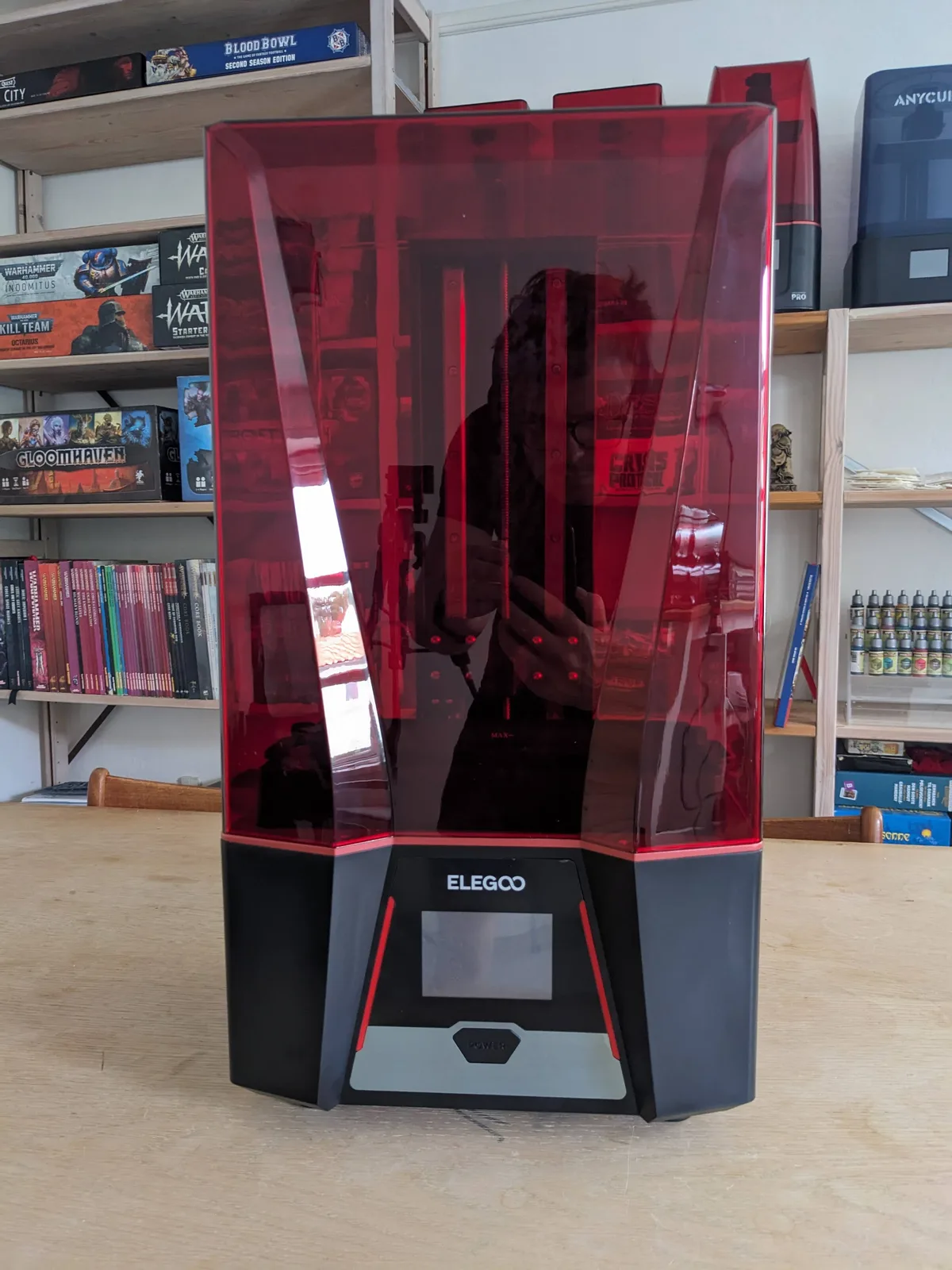 Elegoo Saturn 3 front hero view on a wooden table with the angular hexagonal face, transparent red lid on, and bookshelves behind showing Blood Bowl, Warhammer, and Gloomhaven boxes