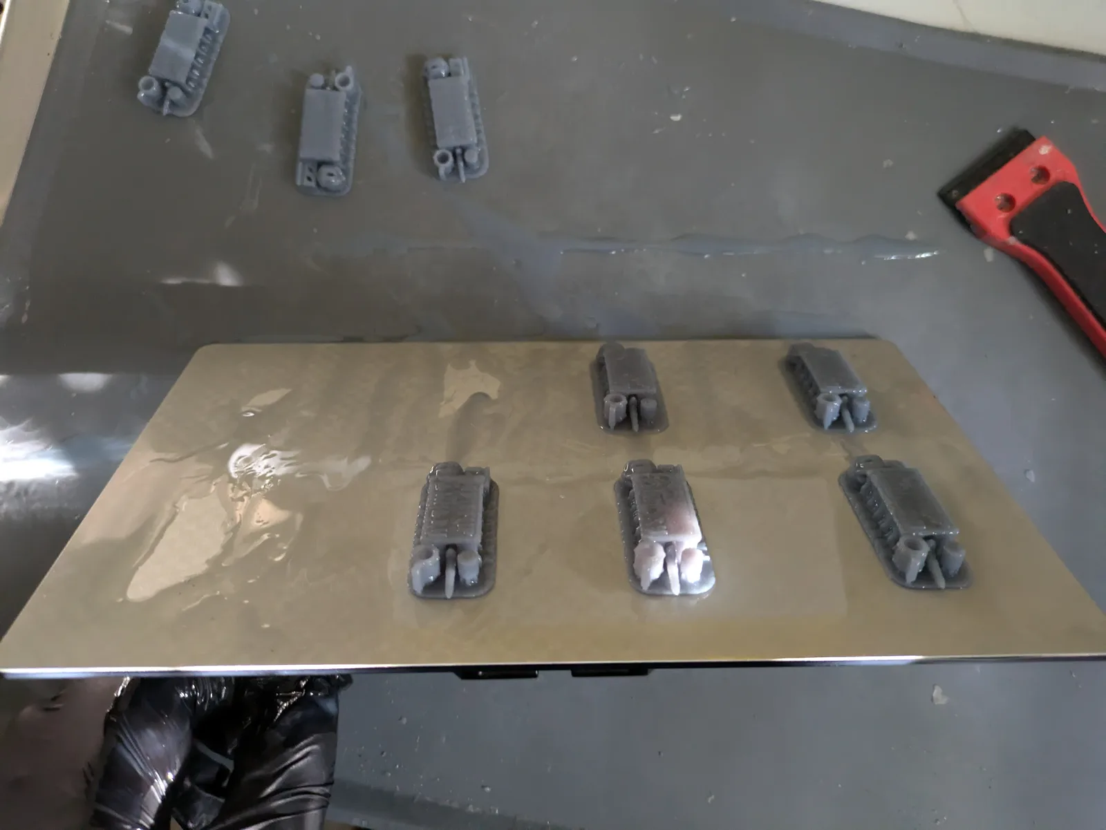 Black gloved hand with a red handled scraper removing six small grey resin prints from a steel build plate