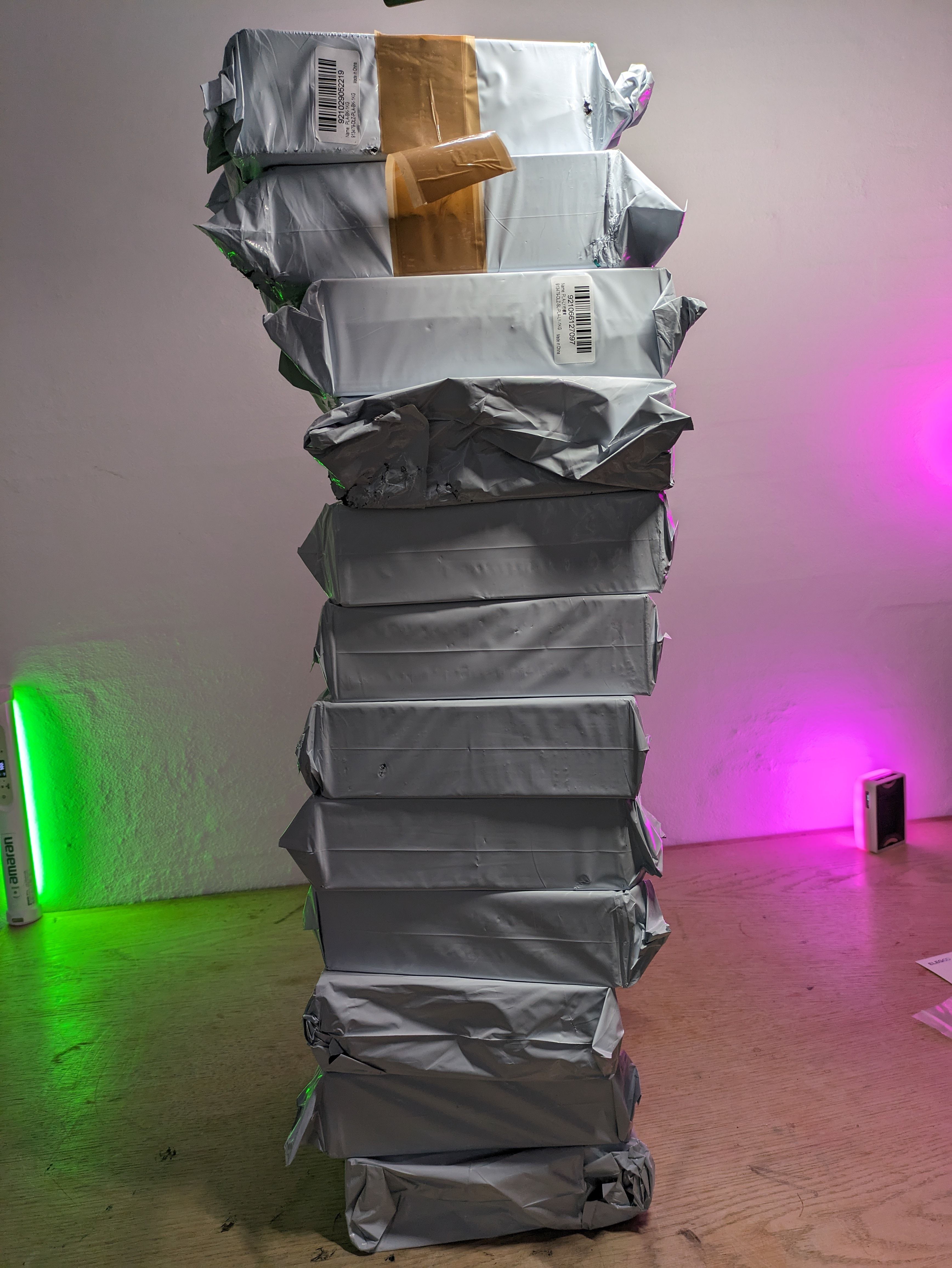 Tall stacked tower of grey Sunlu filament spools wrapped in silver foil against a white wall with green and pink ambient light