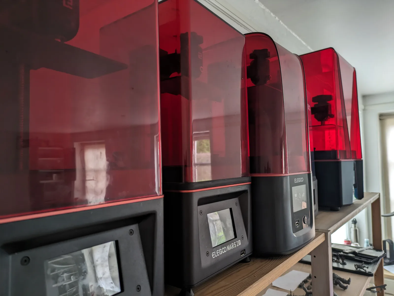 Three Elegoo resin printers in a row on a wooden workshop shelf, all with red acrylic lids, centre unit showing ELEGOO MARS 2B front panel and touchscreen, natural daylight through windows behind