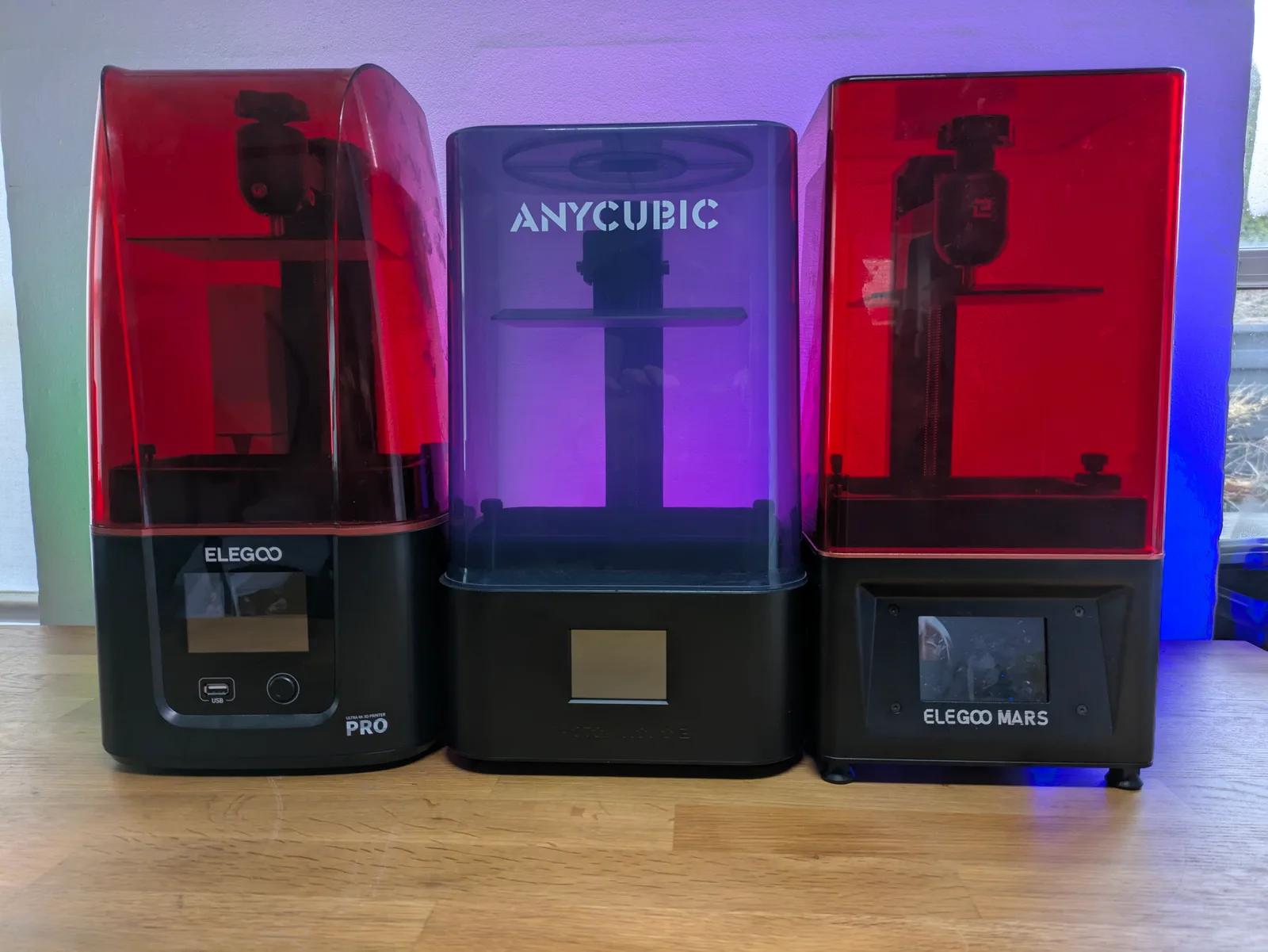 Three resin printers side by side on a wooden surface: an Elegoo with a Pro label on the left, an Anycubic Photon with a glowing transparent hood in the centre, and an Elegoo Mars on the right