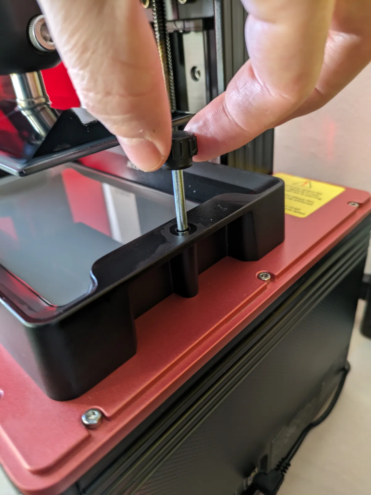 Close-up of a thumb tightening the Mars 4 DLP vat fixing screw with the vat seated on the LCD screen, red printer body and yellow warning sticker visible, detail shot for vat installation