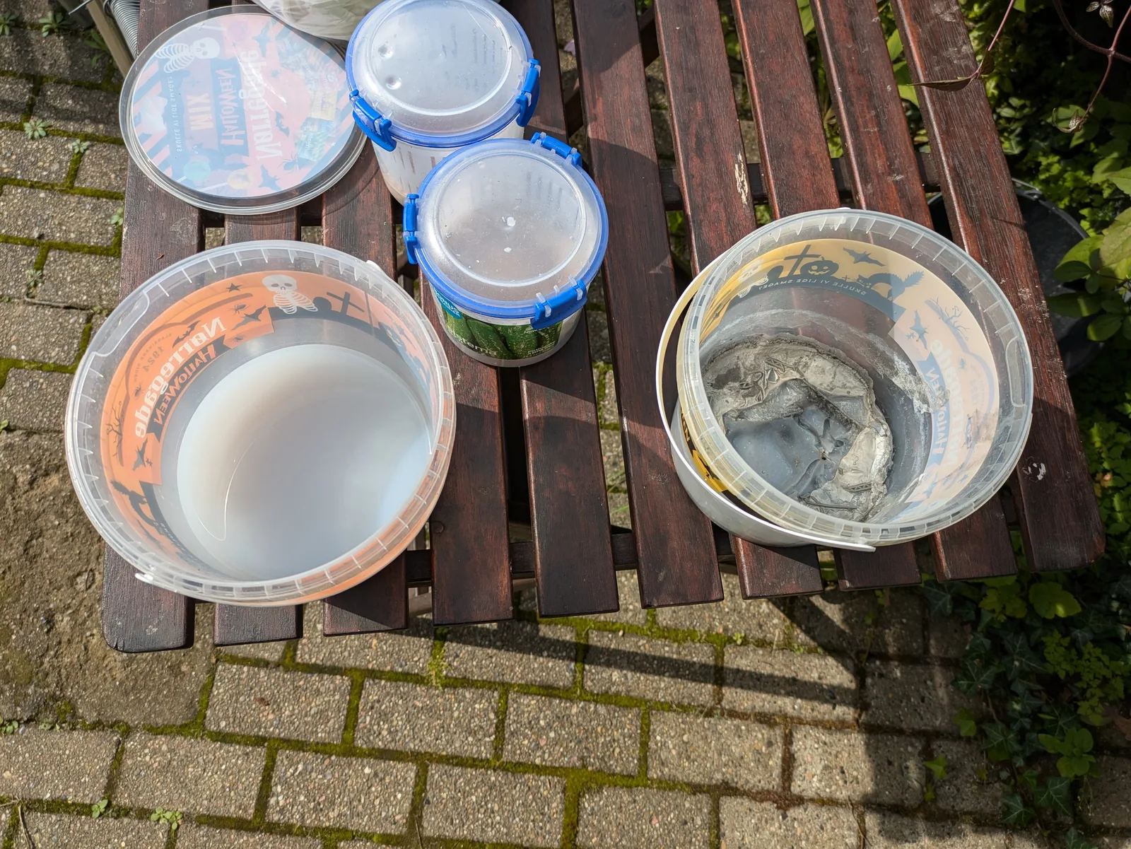 Overhead view of an outdoor wash setup on a wooden garden table, two open bowls side by side, one milky with isopropyl alcohol and one cloudy with grey resin residue