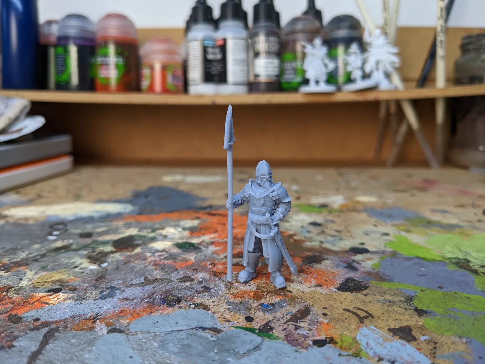 Tall grey resin spear-bearing armoured sentinel standing on a paint stained workbench, finely detailed plate armour and layered garments, long spear held vertically, Army Painter and Citadel paint bottles behind, printed at 35 microns on Mars 3 Pro
