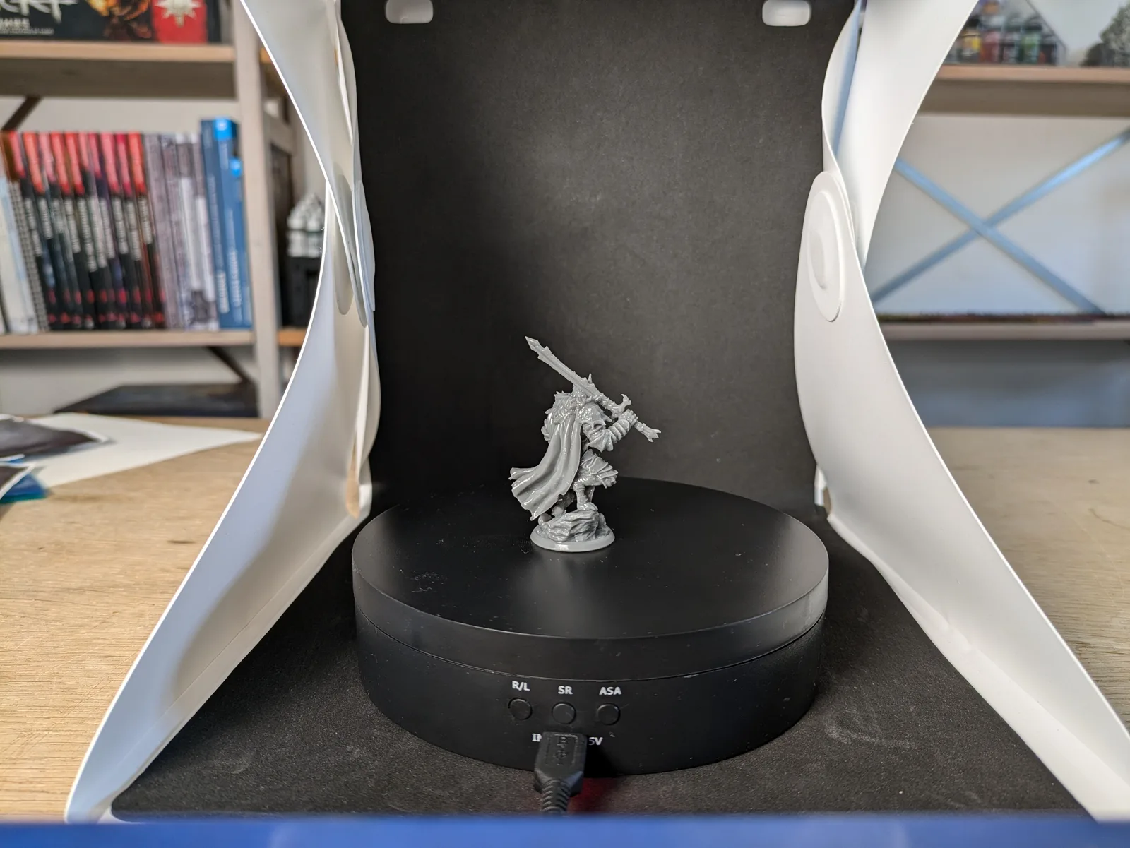 Grey resin dynamic warrior with enormous long sword raised in right hand, left arm extended, flowing cloak and plate and leather armour, on a black rotating turntable in a white and black lightbox