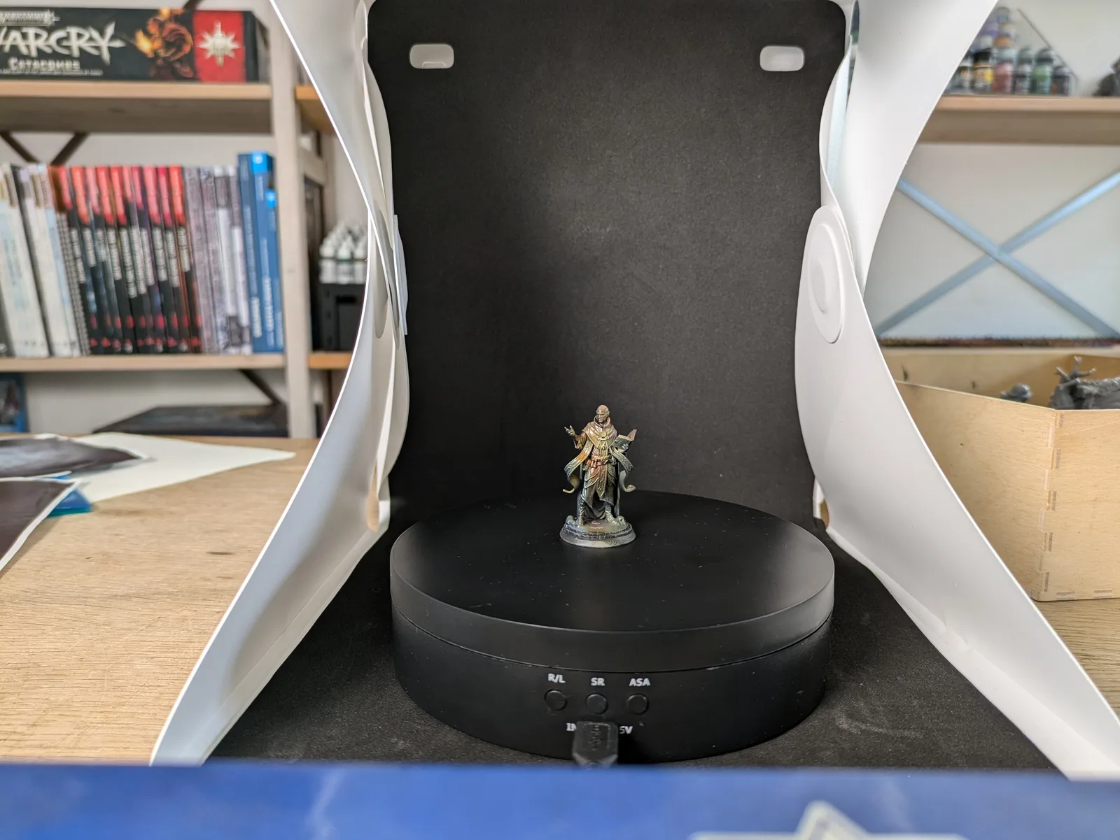 Painted Flesh of Gods Void Envoy character on a turntable in a lightbox, bald robed figure in golden and bronze tones with a dark outer robe, contemplative pose, green-tipped scenic base