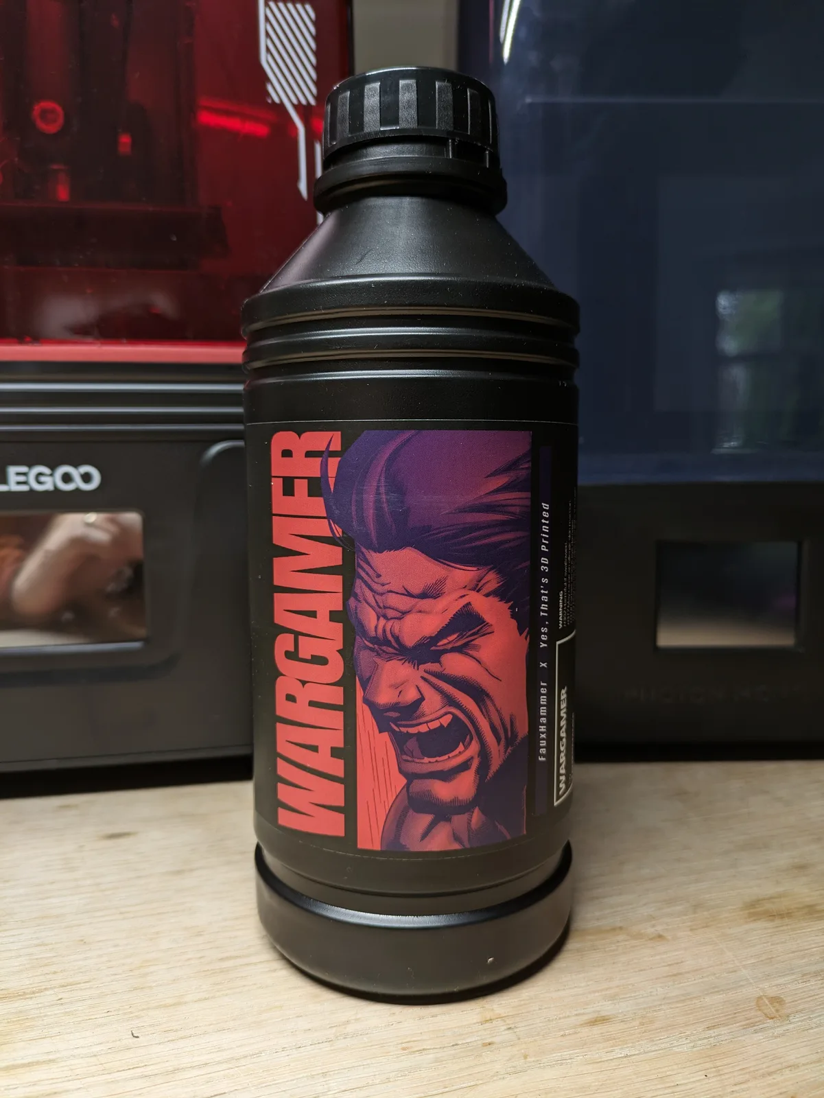 Wargamer Resin bottle on a wooden bench with the red acrylic lid of an Elegoo Saturn behind it, the label readable including FauxHammer X Yes That's 3D Printed