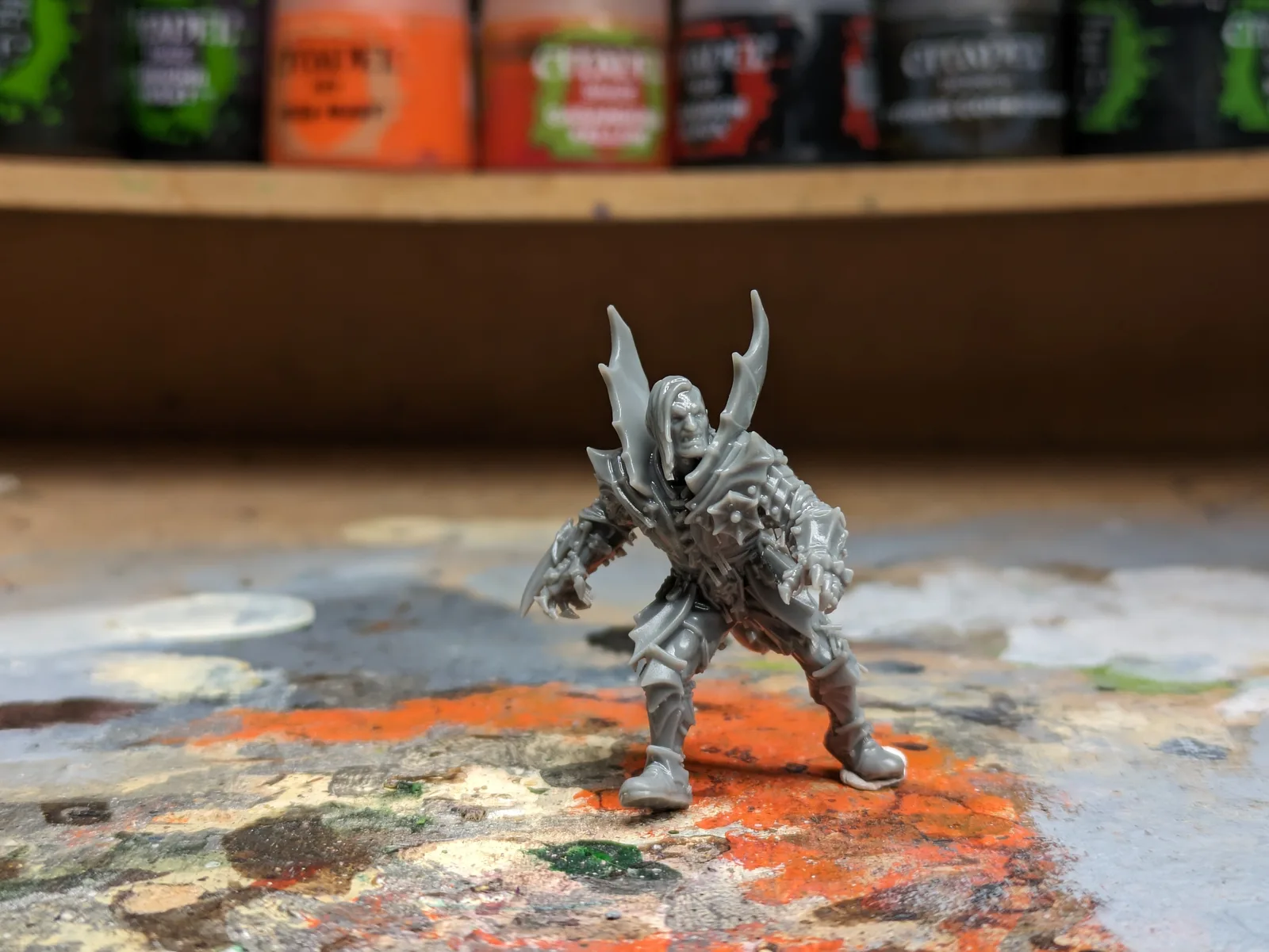 Single grey resin printed dark fantasy warrior with swept back wing armour, posed on a paint palette in front of Citadel paints