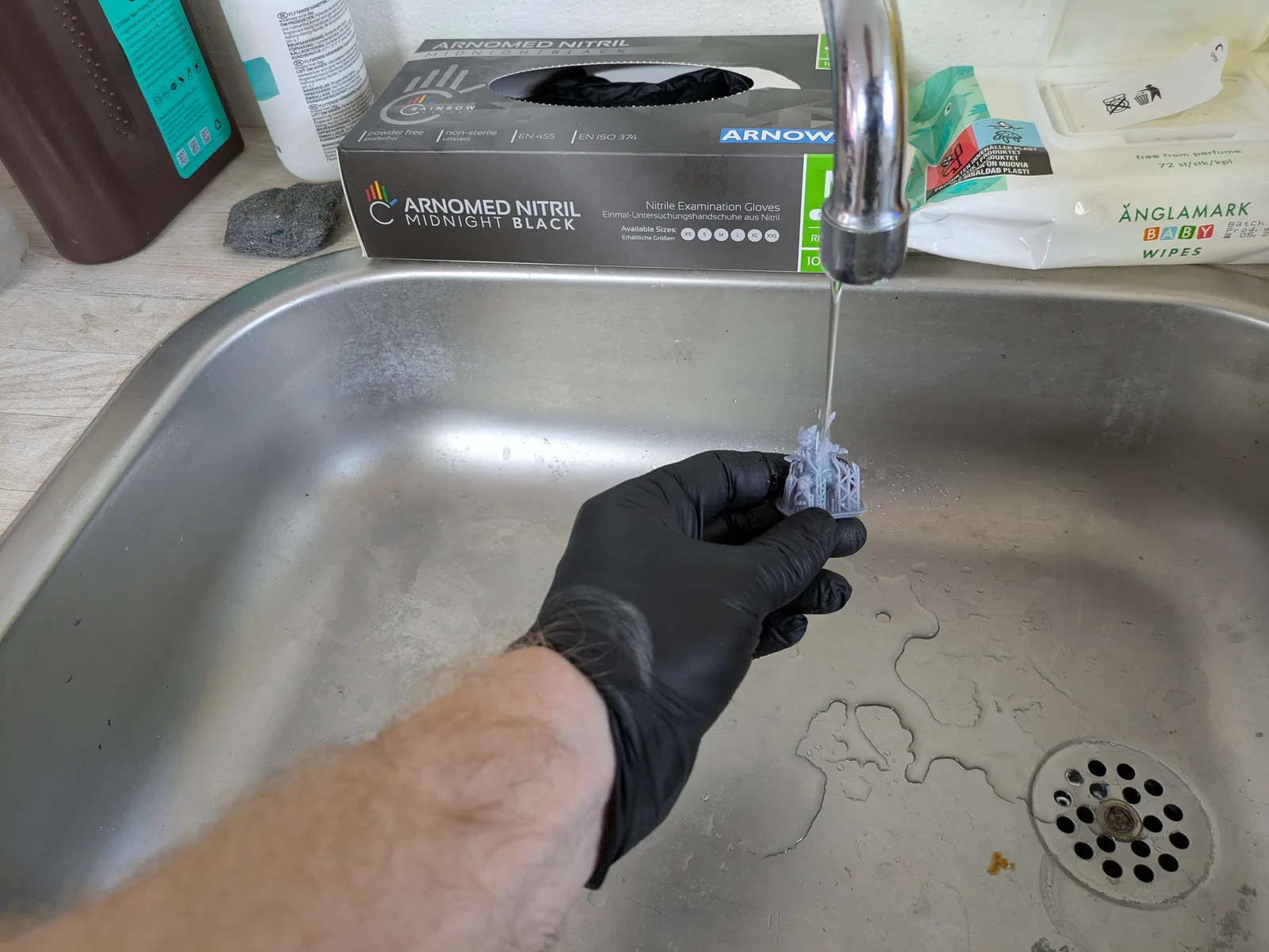 Black nitrile gloved hand holding a small grey resin print under running tap water in a kitchen sink
