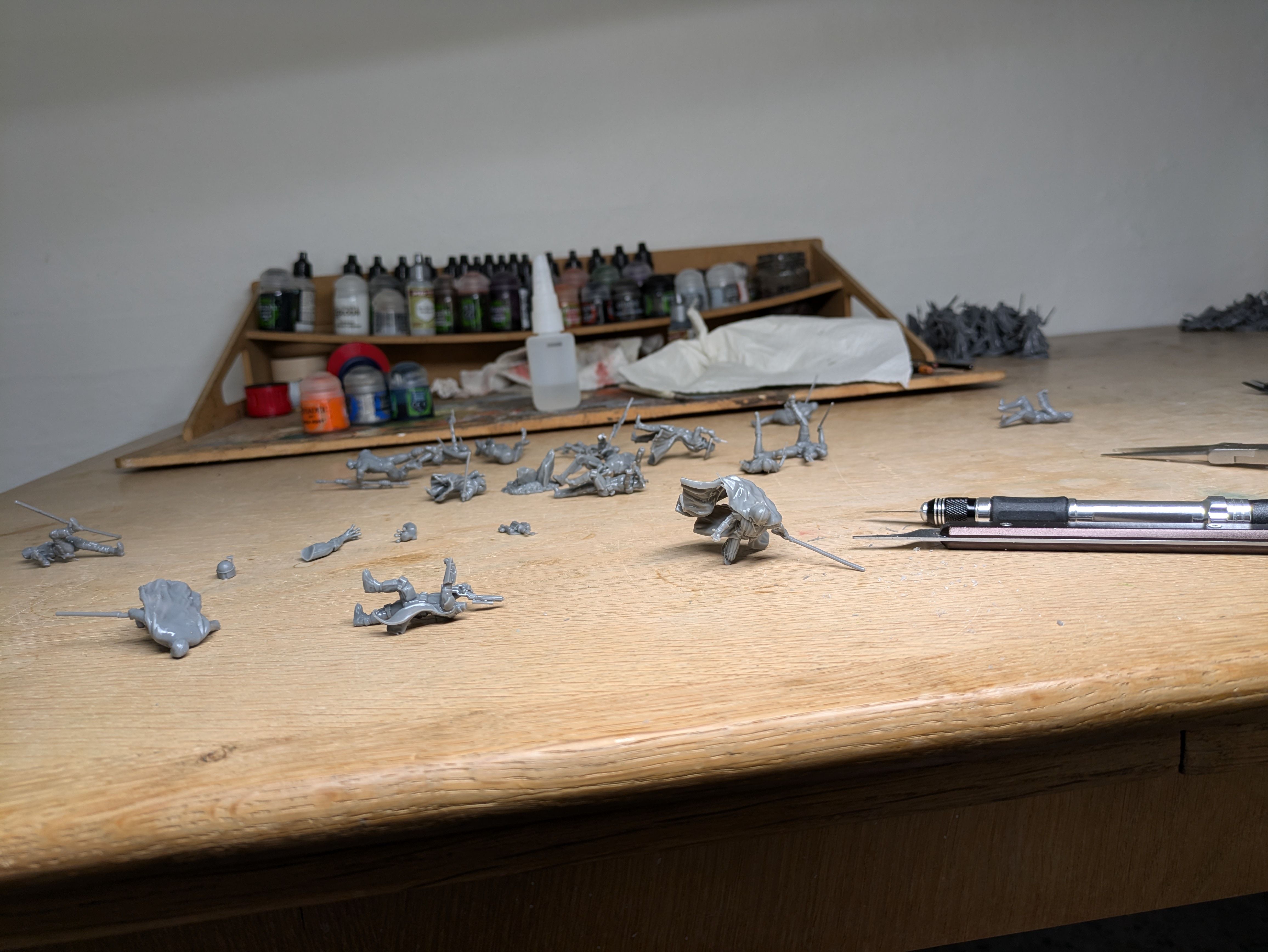 Hobby workbench covered with dozens of unpainted grey resin miniatures after support removal, with hobby tweezers and Army Painter and Citadel paints on the shelf behind